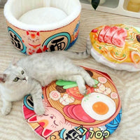 Thumbnail for A white cat lounges near a KittyNook Cat Co. Giant Ramen Bowl Cat Bed and sushi pillow on a colorful mat.