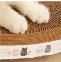 Thumbnail for Cat’s white paws on the KittyNook Cat Co. Oval Corrugated Cat Scratching Board with cute illustrated edges.