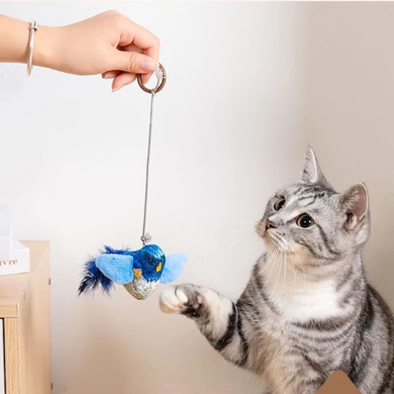 A grey tabby reaches for a KittyNook Cat Co. Ultimate Interactive Chirping Bird Cat Toy held above a wooden table.