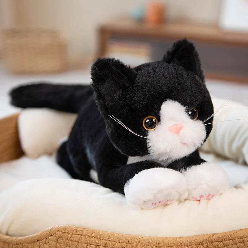 The Snuggle Soft Cat Plush Doll by KittyNook Cat Co. rests on a cream pet bed indoors, showing off its big eyes.
