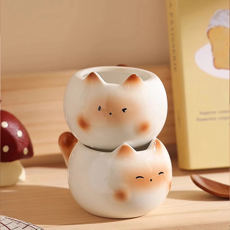 Two Siamese Cat Coffee Cups by KittyNook Cat Co. are stacked on a wooden surface beside a book and decor.