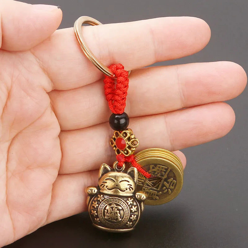 A hand holds the KittyNook Cat Co. Lucky Paws Fortune Cat Keychain with coins and red string against a black background.