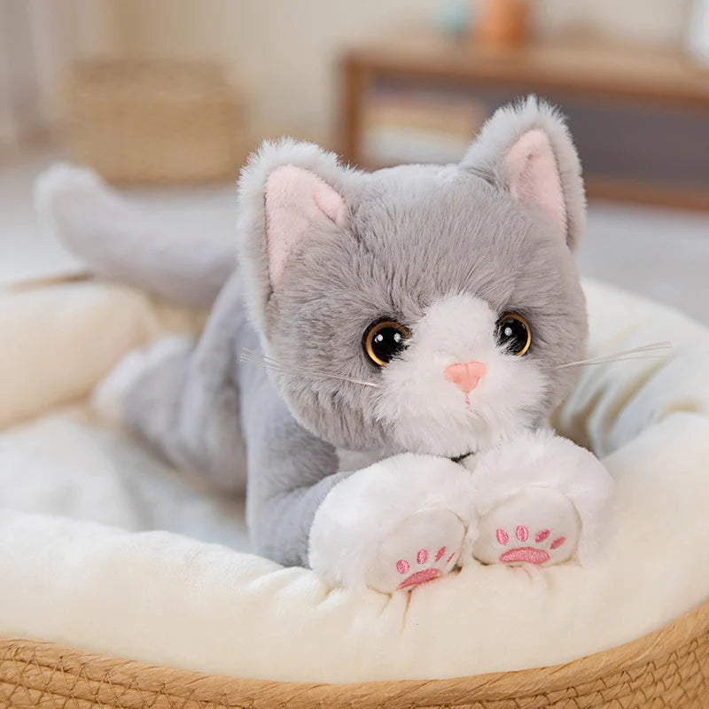 The Snuggle Soft Cat Plush Doll by KittyNook Cat Co. rests in a cozy, cream-colored pet bed with big eyes.