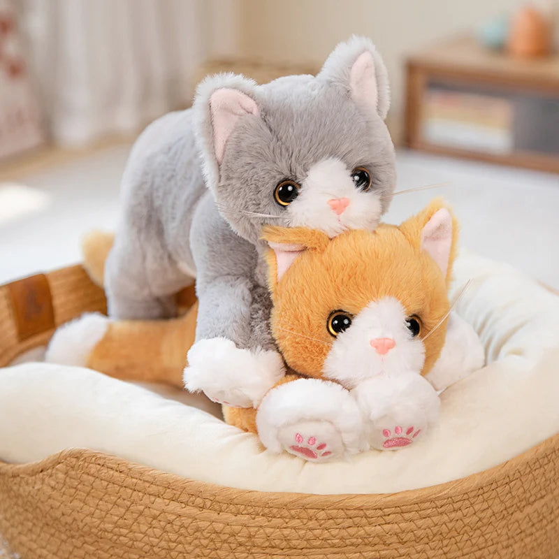 Two Snuggle Soft Cat Plush Dolls by KittyNook Cat Co., one gray and one orange, cuddle on a round pet bed indoors.