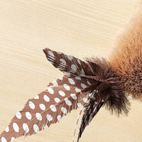 Thumbnail for KittyNook Cat Co. Squeaky Plush Cat Toy in tan with brown feathers and white spots on a light wooden surface.