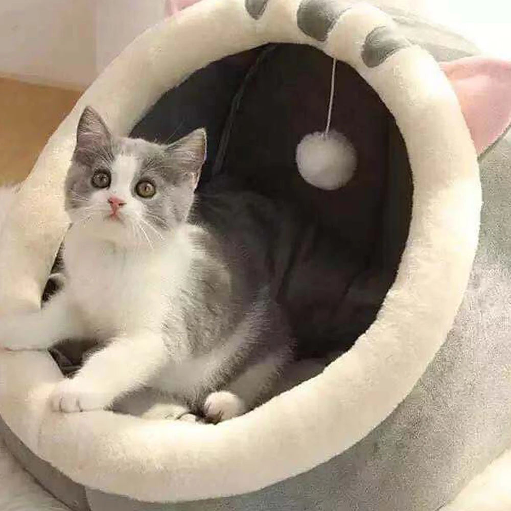 A gray and white cat relaxes in the KittyNook Cat Co. Cute Comfortable Cat Nest Bed with a hanging pom-pom toy.