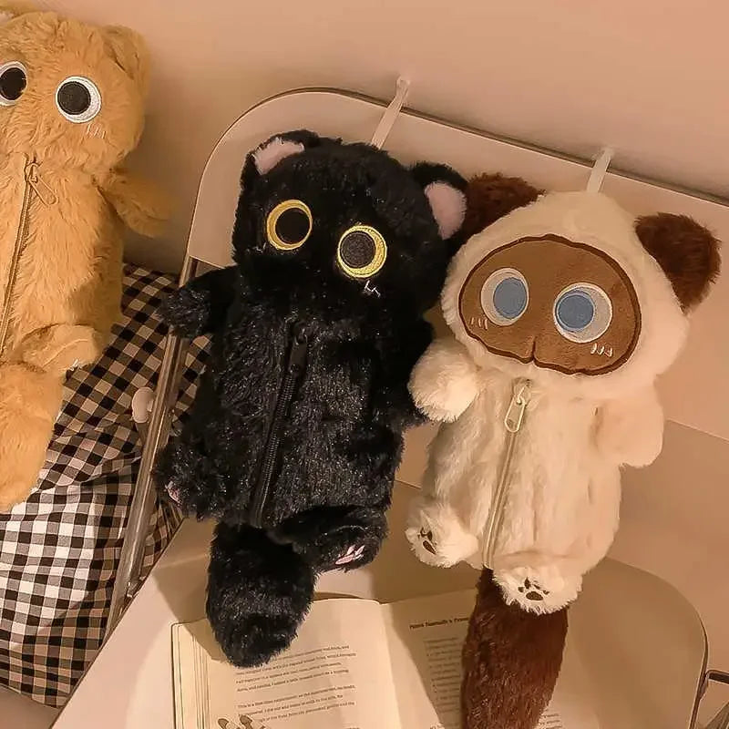 Two plush toys with large eyes sit on a desk near a book, plaid bag, and KittyNook Cat Co. Kawaii Big-Eyed Cat Plush Pencil Case.