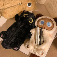 Thumbnail for Three plush pencil cases, including the KittyNook Cat Co. Kawaii Big-Eyed Cat Plush Pencil Case, lie by an open book.