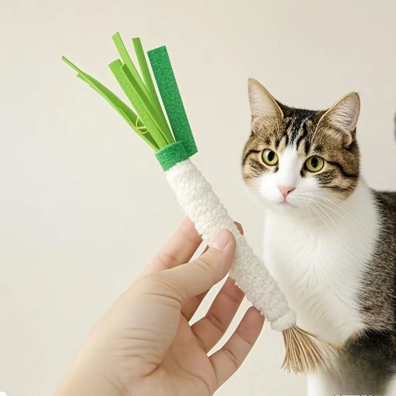 A hand holds KittyNook Cat Co.'s Ultimate Kitten Teething Wand Toy as a cat curiously watches in the background.