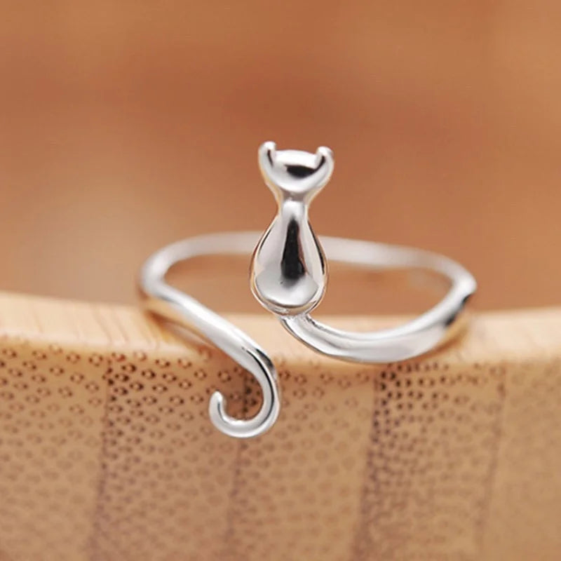The Cute Adjustable Silver Cat Ring by KittyNook Cat Co. features a curled tail and is shown on a wooden surface.