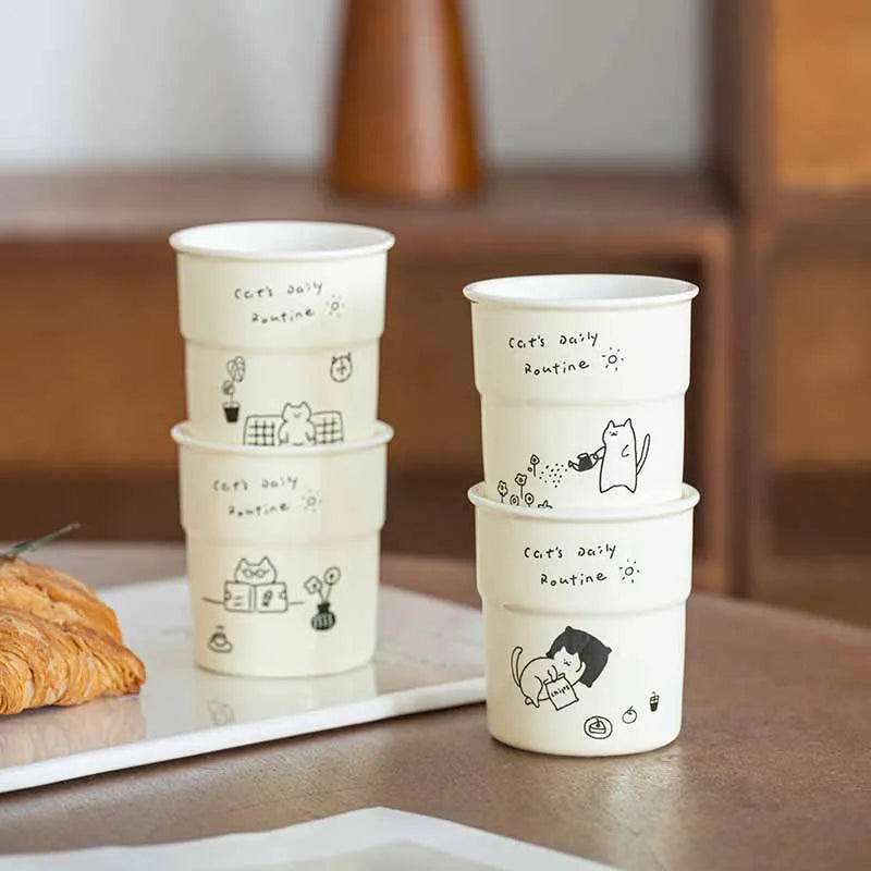 Six KittyNook Cat Co. Cute Illustrated Ceramic Cat Mugs, white with "Cat's Daily Routine" text, beside a croissant.