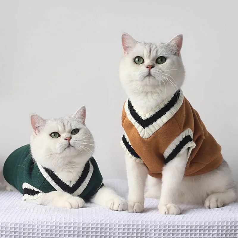 Two white cats wear green and brown Cozy Fluff Cat Sweaters by KittyNook Cat Co., sitting side by side on a white surface.