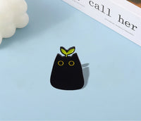 Thumbnail for KittyNook Cat Co. Black Cat Enamel Brooch with yellow eyes and green sprout, shown on blue near a book.