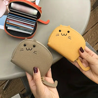Thumbnail for Two hands hold KittyNook Cat Co. Cute Multi-Card Cat Purses—one gray, one yellow—near an open wallet on a table.