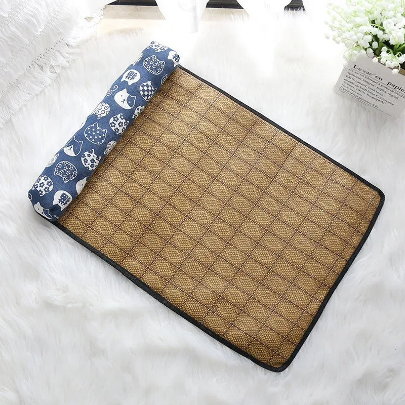 The KittyNook Cat Co. Natural Rattan Cat Sleeping Mat with a blue-white pillow rests on a fluffy surface near a plant.