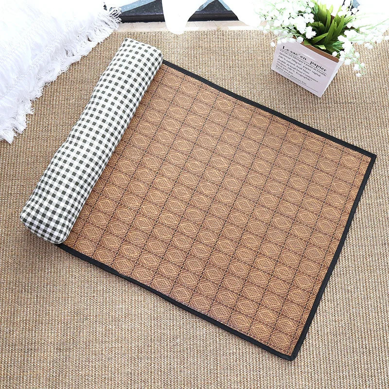 The KittyNook Cat Co. Natural Rattan Cat Sleeping Mat with checkered pillow sits on a woven carpet by a flower pot.