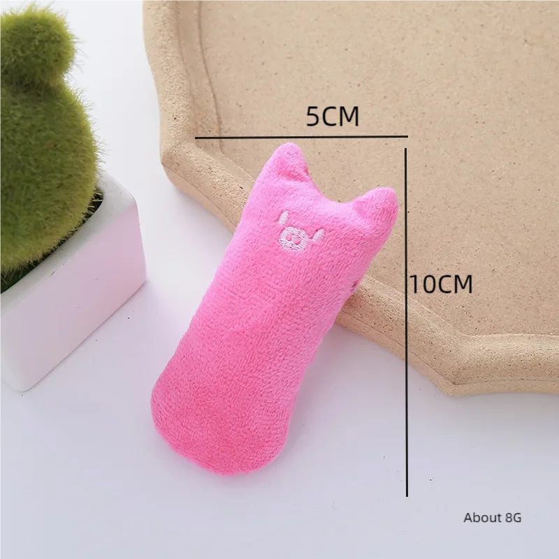 The Safe Catnip Chew Toy by KittyNook Cat Co., 10x5cm, pink with embroidered face, is on a beige tray by a green plant.