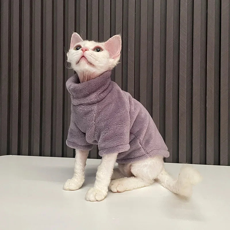 A hairless kitten in a KittyNook Cat Co. warm winter hoodie sits on a table against a striped gray background.