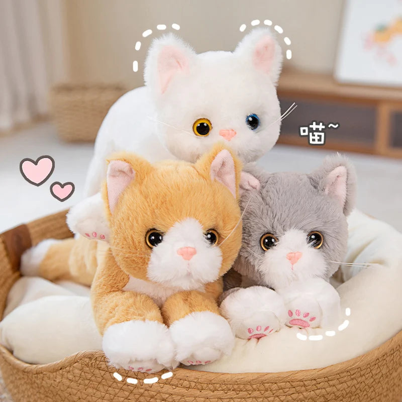 Three Snuggle Soft Cat Plush Dolls by KittyNook Cat Co.—white, orange & gray—sit together on a heart-decorated cushion.