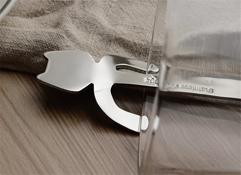 The Cat Cling Stainless Steel Mug Spoon by KittyNook Cat Co. peeks out from under a glass on beige fabric over wood.