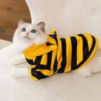 Thumbnail for A white cat in the KittyNook Cat Co. Cute Bee Costume lies on a fuzzy surface, looking up.