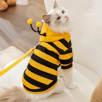 Thumbnail for Fluffy white cat in KittyNook Cat Co. Cute Bee Costume, with antennae, sits on a chair looking up.